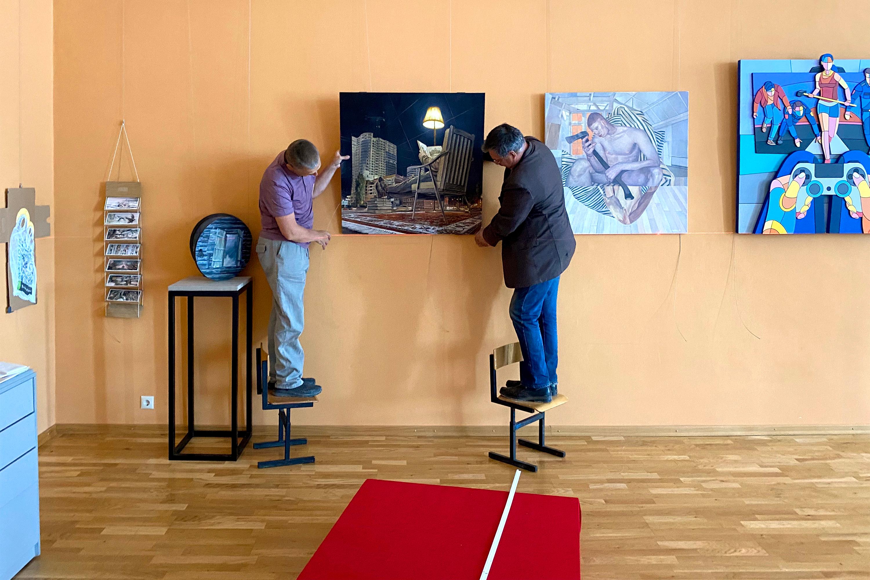 Второй день монтажа (развесочный). Специально обученные люди выравнивают по лазерному уровню картину Андрея Оленева, подвешенную на рельсе с помощью тросов